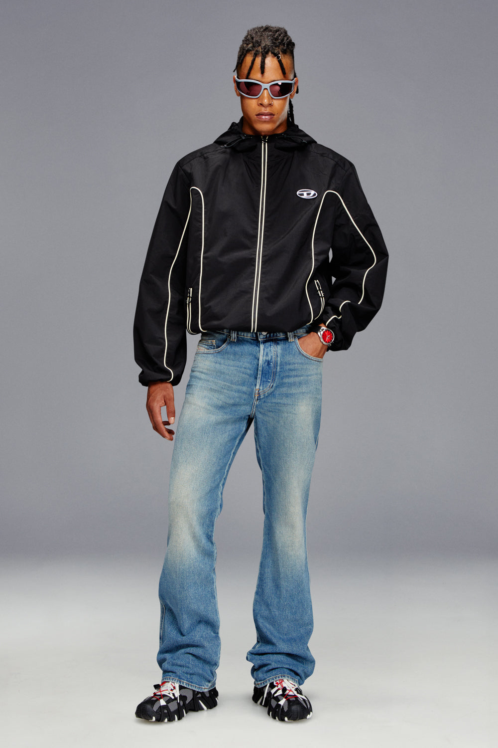 Person wearing a black jacket with a logo, blue jeans, and sunglasses on a gray background
