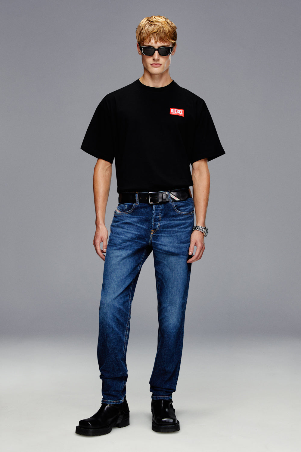 Man wearing a black t-shirt with a red logo, blue jeans, and black shoes on a gray background