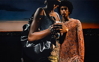 Two models wearing edgy, metallic and textured avant-garde fashion against a twilight skyline.