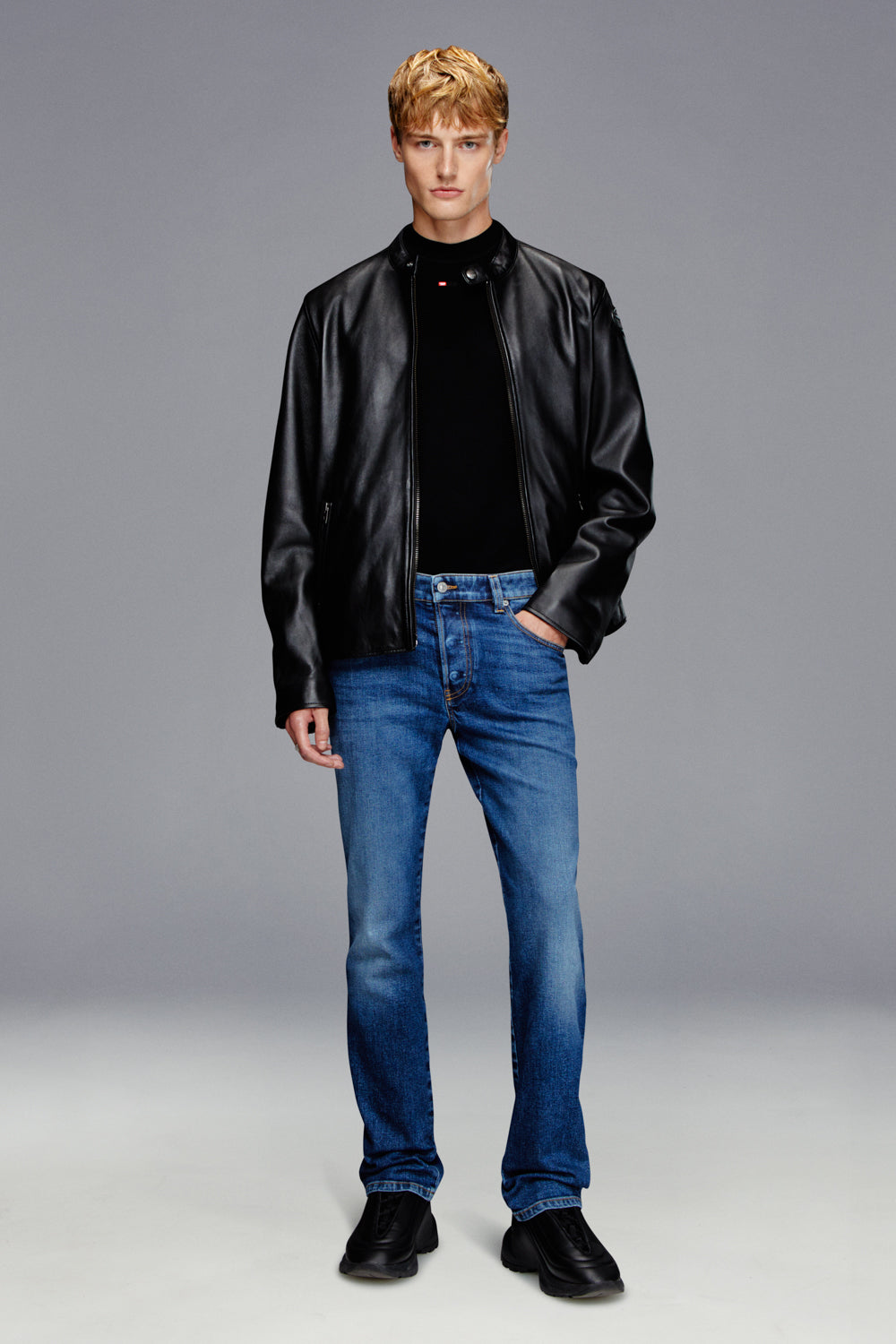 Man wearing a black leather jacket, black shirt, and blue jeans on a gray background